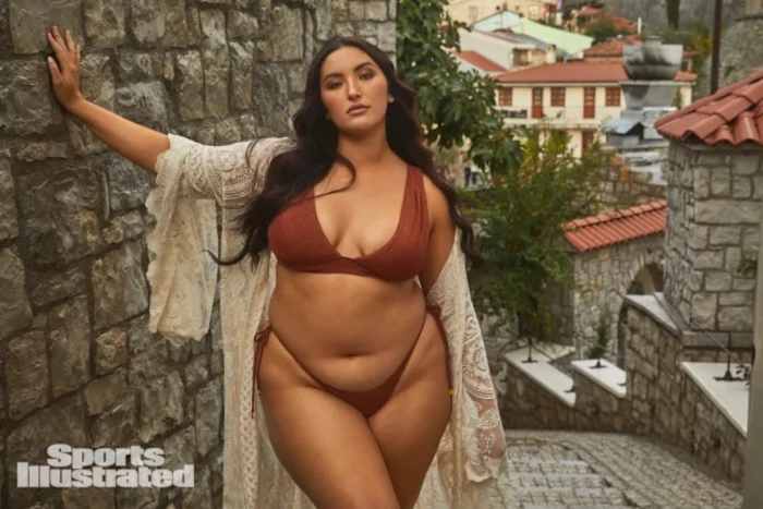 Yumi Nu stands on the cobblestone street and leans against a stone wall, wearing a deep rust-colored bikini and a white lace coverup.